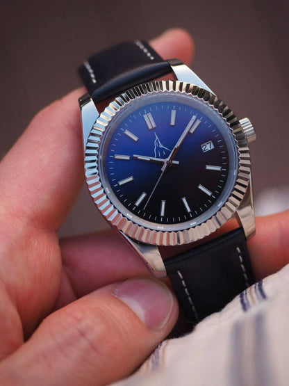 Gros plan de la montre L'Aiguille d'Étretat tenue en main, mettant en valeur le logo discret et la lunette cannelée iconique en acier. Vue détaillée du cadran bleu dégradé, des index lumineux, des aiguilles dauphines et du guichet dateur sur le bracelet cuir noir.