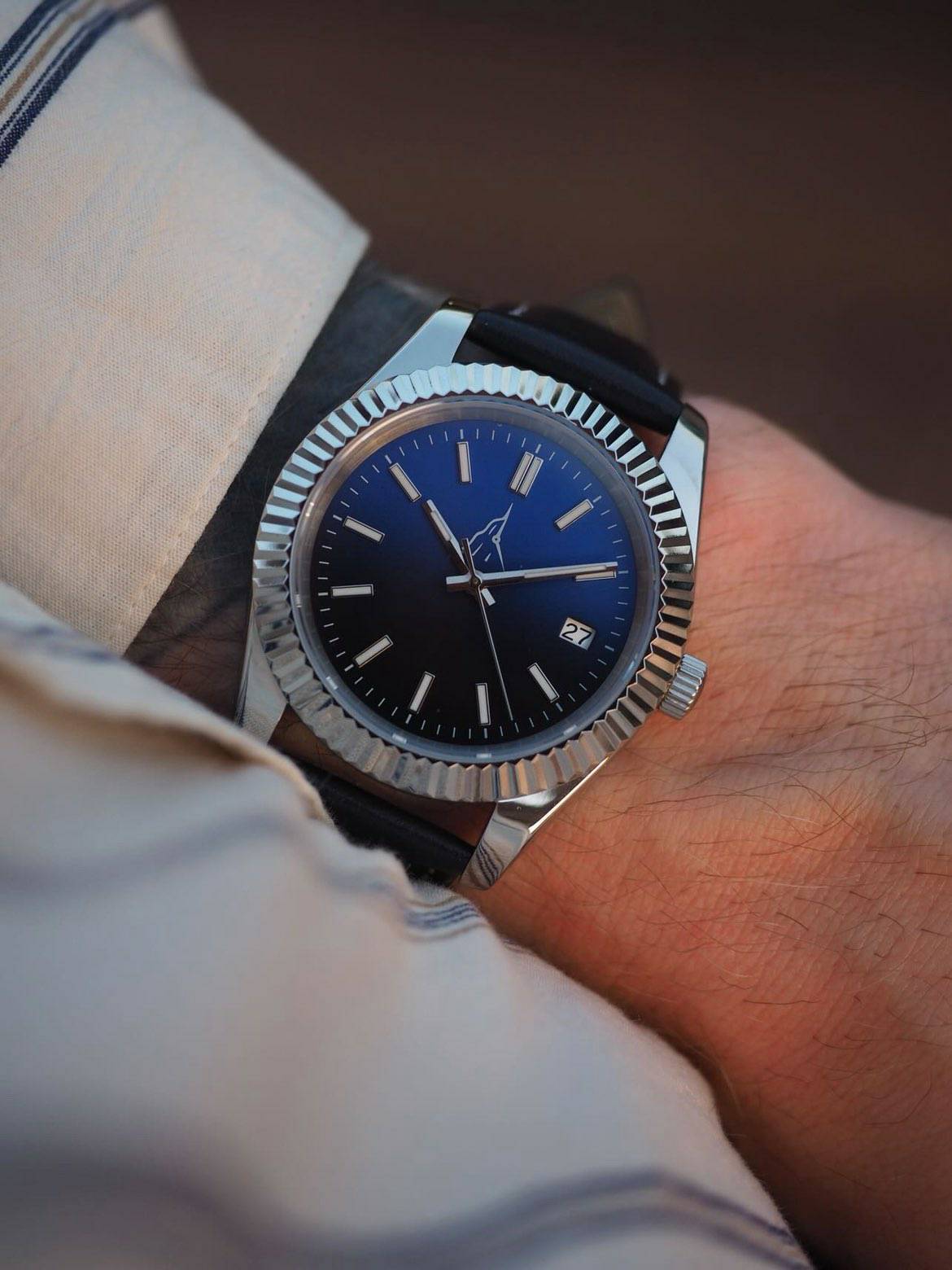 Montre automatique premium L'Aiguille d'Étretat portée au poignet, cadran bleu dégradé soleillé, lunette cannelée en acier chirurgical poli, guichet date, bracelet en cuir noir piqué main, style élégant avec chemise rayée.